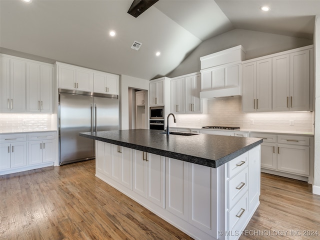 kitchen featuring vaulted ceiling, sink, built in appliances, white cabinets, and an island with sink