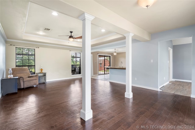 unfurnished living room with a wealth of natural light, dark wood-type flooring, ornate columns, ceiling fan, and a raised ceiling