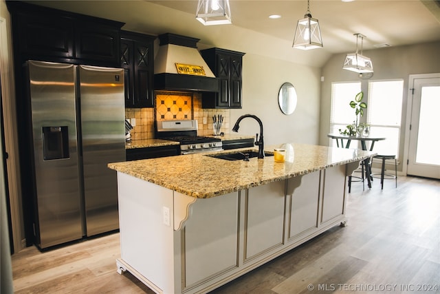 kitchen with an island with sink, stainless steel appliances, decorative light fixtures, premium range hood, and backsplash