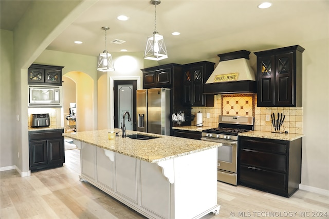 kitchen with stainless steel appliances, hanging light fixtures, a kitchen island with sink, custom exhaust hood, and light wood-type flooring