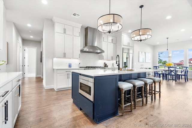 kitchen featuring white cabinetry, wall chimney range hood, built in appliances, hanging light fixtures, and a center island with sink