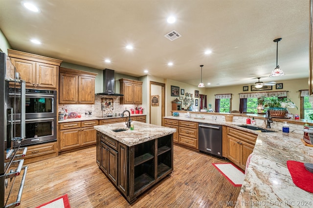 kitchen featuring hanging light fixtures, light hardwood / wood-style floors, stainless steel appliances, a center island with sink, and wall chimney range hood