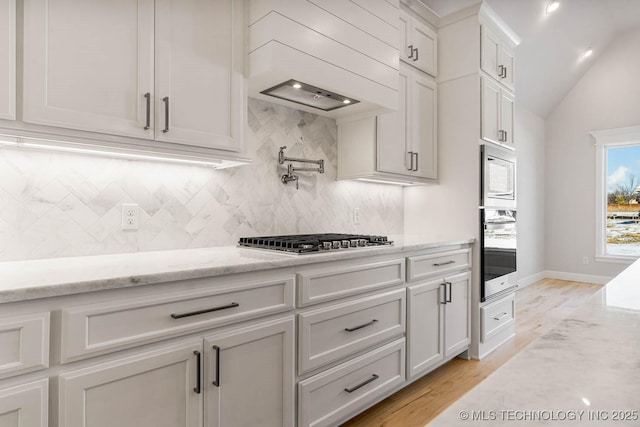 kitchen featuring vaulted ceiling, appliances with stainless steel finishes, white cabinets, light stone counters, and custom range hood