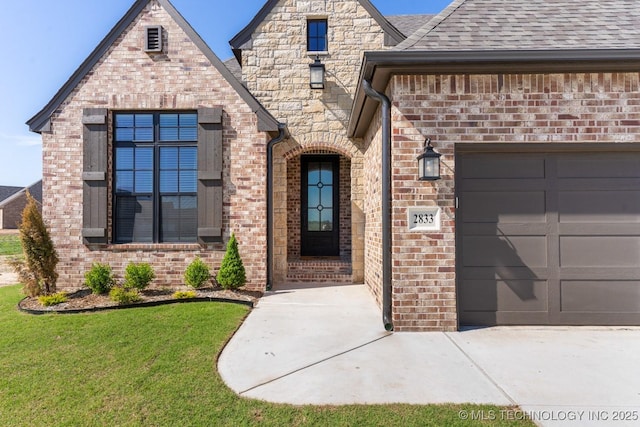 view of exterior entry featuring roof with shingles, an attached garage, brick siding, a lawn, and driveway