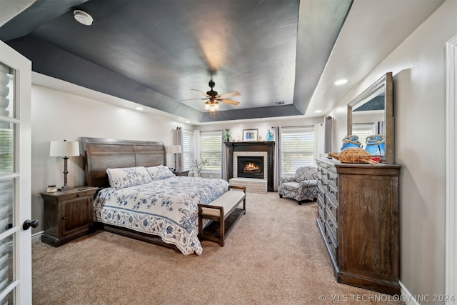 bedroom with a brick fireplace, ceiling fan, a tray ceiling, and light carpet