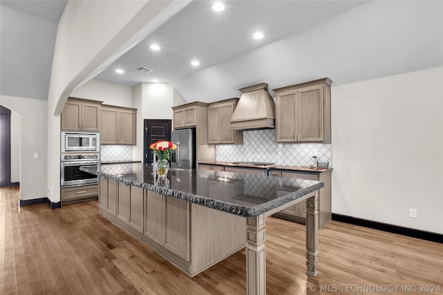 kitchen with appliances with stainless steel finishes, a kitchen island, decorative backsplash, light wood-type flooring, and custom exhaust hood