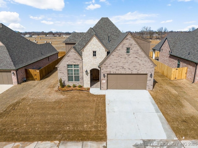 french country inspired facade featuring a shingled roof, fence, concrete driveway, and brick siding