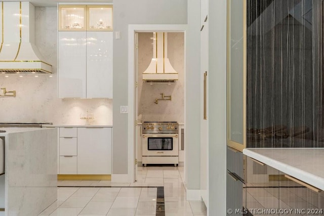 kitchen featuring tasteful backsplash, stainless steel stove, custom range hood, light tile patterned floors, and white cabinets