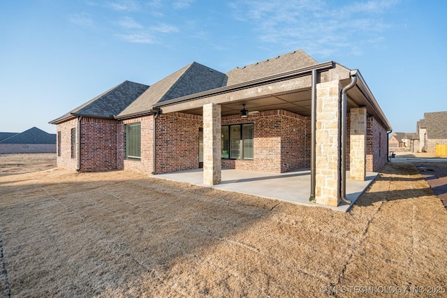 rear view of house with ceiling fan, brick siding, stone siding, roof with shingles, and a patio area