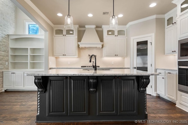 kitchen featuring dark hardwood / wood-style flooring, premium range hood, sink, a kitchen island with sink, and light stone counters