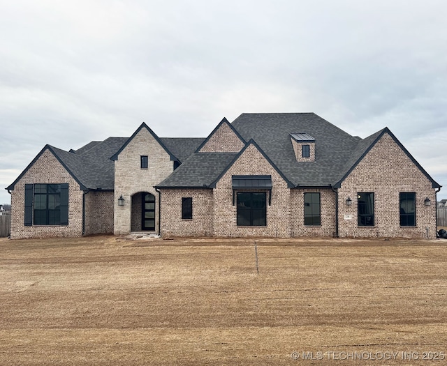 french country home featuring roof with shingles, a front yard, and brick siding