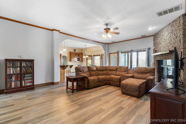 living room with ceiling fan with notable chandelier, light wood-type flooring, crown molding, and a stone fireplace