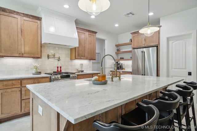 kitchen with a kitchen bar, an island with sink, hanging light fixtures, stainless steel appliances, and premium range hood