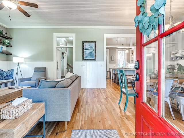 living area with a ceiling fan, a wainscoted wall, crown molding, and light wood-style flooring