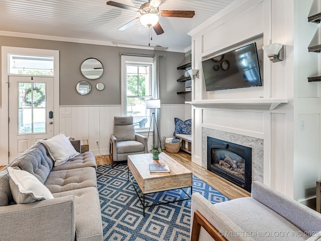 living room with a premium fireplace, ornamental molding, a ceiling fan, wainscoting, and wood finished floors