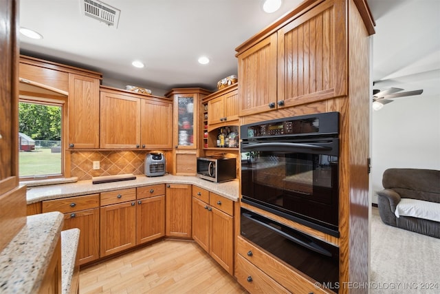 kitchen with black oven, decorative backsplash, ceiling fan, light hardwood / wood-style floors, and light stone countertops