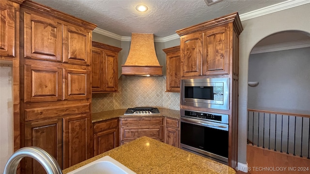 kitchen featuring decorative backsplash, light stone countertops, ornamental molding, custom range hood, and stainless steel appliances