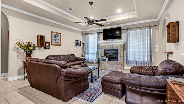 living room with ceiling fan, a tray ceiling, ornamental molding, a tiled fireplace, and light tile patterned floors