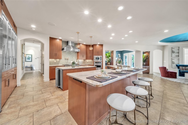 kitchen with arched walkways, a large island, wall chimney range hood, appliances with stainless steel finishes, and backsplash