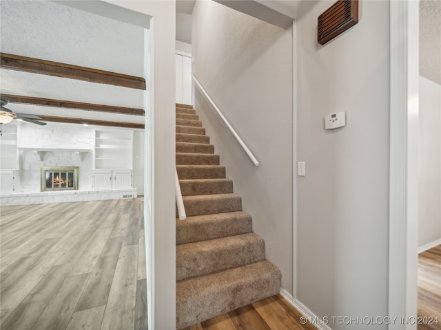 staircase with a fireplace, hardwood / wood-style floors, ceiling fan, and beamed ceiling