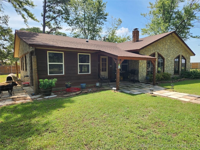 back of house featuring a shingled roof, a lawn, stone siding, a chimney, and a patio area