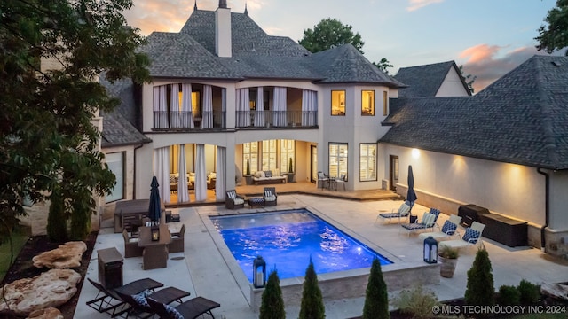 back house at dusk featuring a balcony, a patio area, french doors, and a swimming pool with hot tub