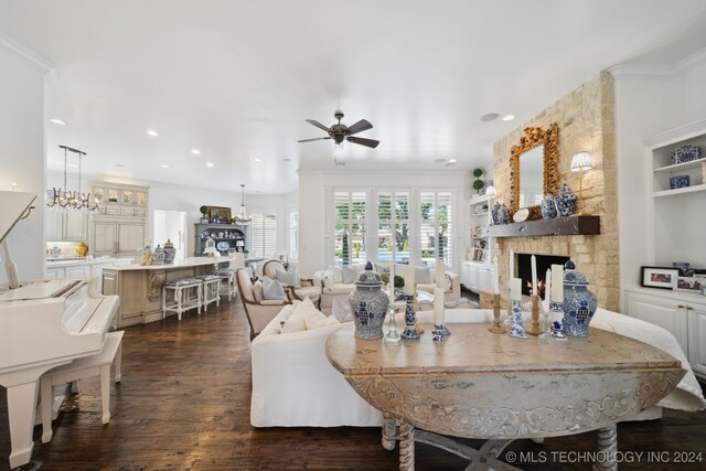 living room with built in features, dark hardwood / wood-style floors, a stone fireplace, ceiling fan with notable chandelier, and crown molding