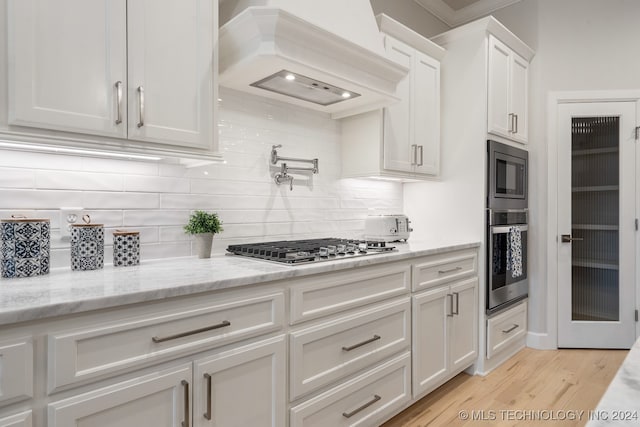 kitchen with backsplash, custom range hood, light wood-type flooring, appliances with stainless steel finishes, and white cabinets