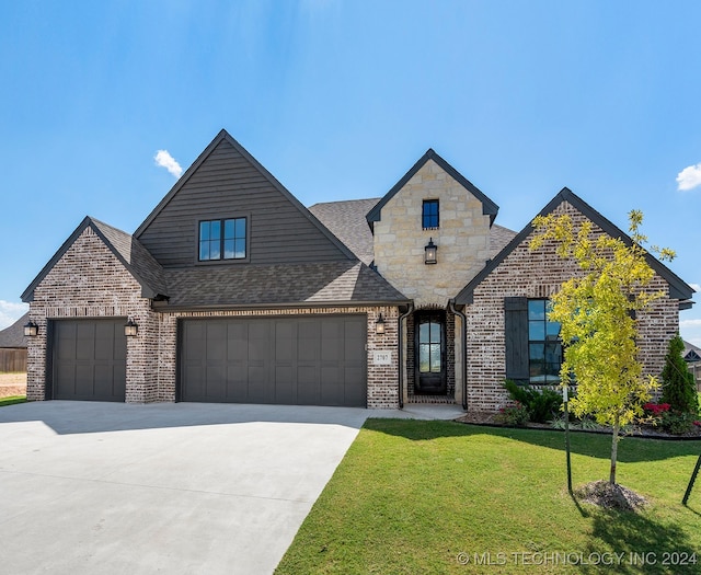 french country inspired facade with a shingled roof, a front yard, driveway, brick siding, and an attached garage