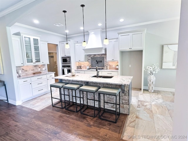 kitchen featuring light stone counters, a center island with sink, custom exhaust hood, stainless steel appliances, and white cabinets