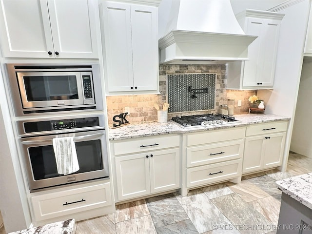 kitchen with light stone countertops, premium range hood, white cabinetry, appliances with stainless steel finishes, and decorative backsplash