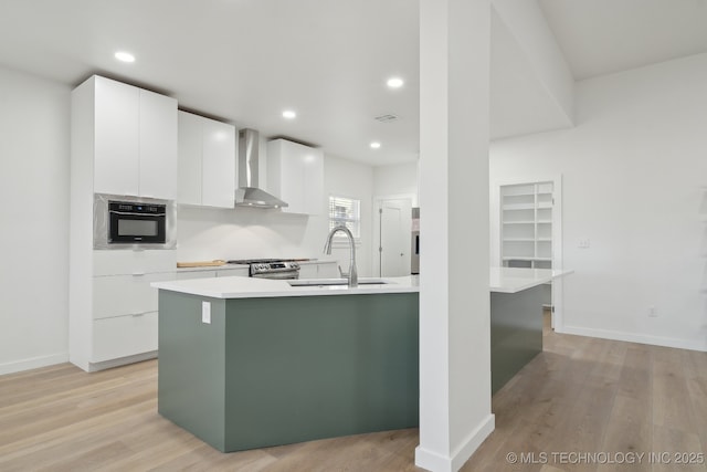 kitchen featuring wall chimney range hood, sink, white cabinetry, stainless steel gas range oven, and a center island with sink