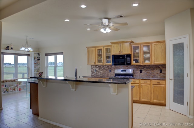 kitchen with dark stone countertops, ceiling fan with notable chandelier, stainless steel range, an island with sink, and a breakfast bar