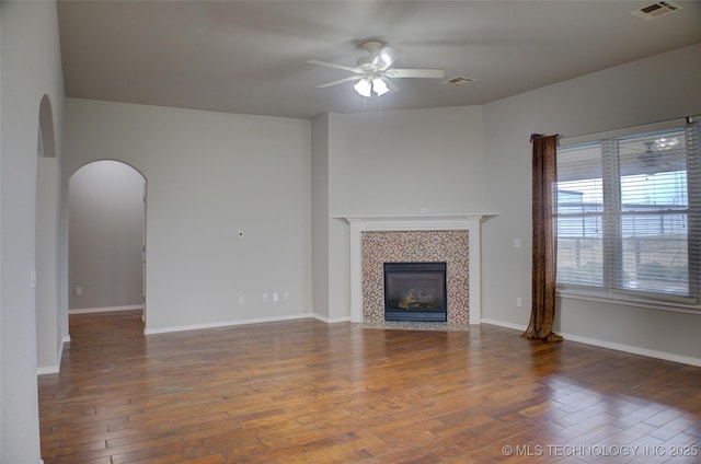 unfurnished living room featuring arched walkways, ceiling fan, a tile fireplace, visible vents, and hardwood / wood-style floors