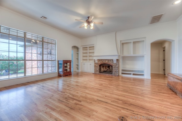 unfurnished living room featuring a stone fireplace, ornamental molding, built in shelves, light wood-type flooring, and ceiling fan