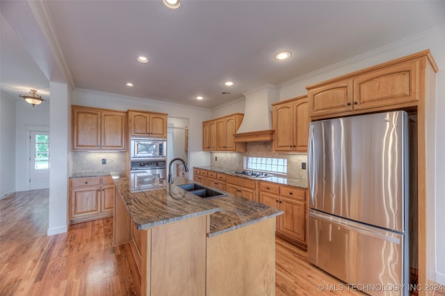 kitchen featuring appliances with stainless steel finishes, premium range hood, sink, light wood-type flooring, and a kitchen island with sink