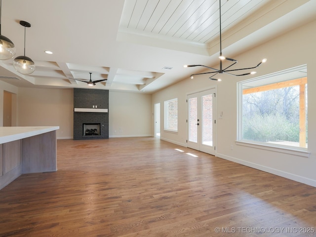 unfurnished living room featuring baseboards, coffered ceiling, wood finished floors, a brick fireplace, and ceiling fan with notable chandelier