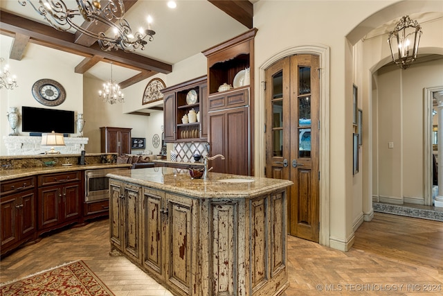 kitchen with sink, decorative light fixtures, beam ceiling, light stone counters, and an island with sink