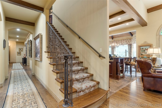 stairs featuring beam ceiling, wood-type flooring, and an inviting chandelier