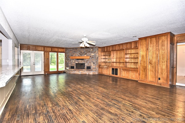 unfurnished living room with built in shelves, ceiling fan, a textured ceiling, a stone fireplace, and dark wood-type flooring