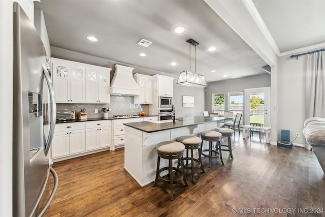 kitchen featuring appliances with stainless steel finishes, a kitchen island with sink, white cabinets, decorative light fixtures, and custom exhaust hood