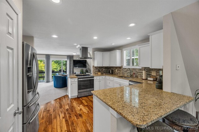 kitchen featuring kitchen peninsula, stainless steel appliances, white cabinetry, and wall chimney range hood