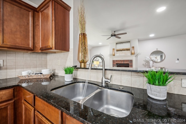 kitchen featuring tasteful backsplash, dark stone counters, ceiling fan, sink, and a fireplace