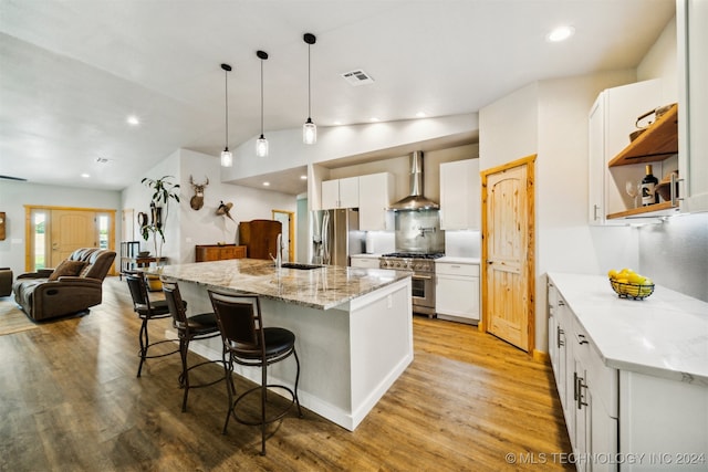 kitchen with white cabinets, appliances with stainless steel finishes, light stone counters, wall chimney range hood, and a center island with sink