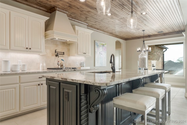 kitchen with a kitchen island with sink, custom exhaust hood, decorative light fixtures, light stone counters, and wood ceiling