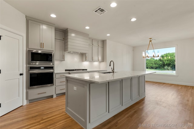 kitchen with a kitchen island with sink, light wood-type flooring, an inviting chandelier, sink, and appliances with stainless steel finishes