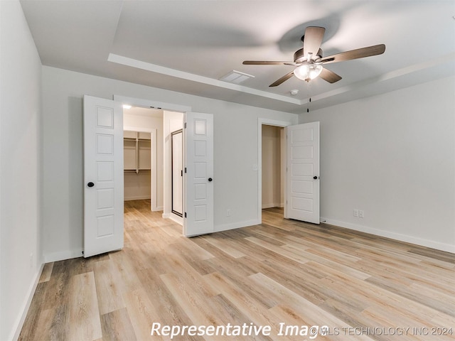 unfurnished bedroom with a spacious closet, a closet, a tray ceiling, light hardwood / wood-style floors, and ceiling fan