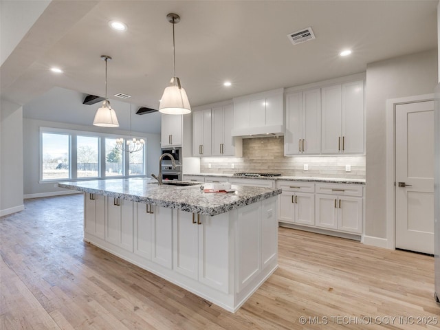 kitchen with white cabinetry, decorative light fixtures, a kitchen island with sink, and light stone countertops