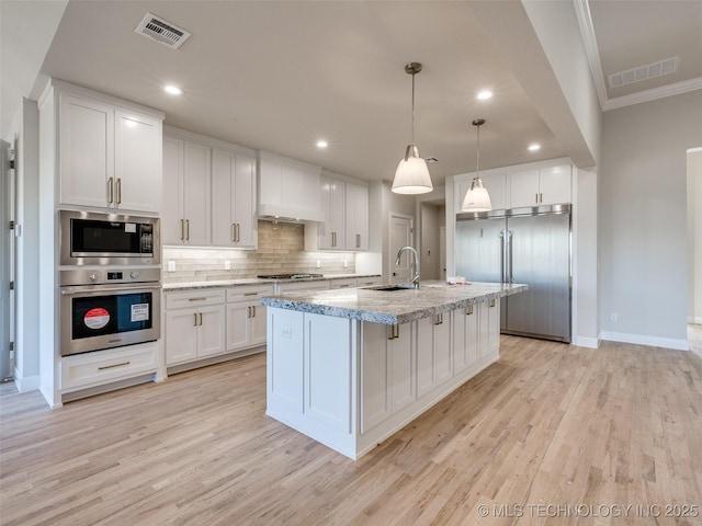 kitchen featuring sink, decorative light fixtures, built in appliances, a kitchen island with sink, and white cabinets