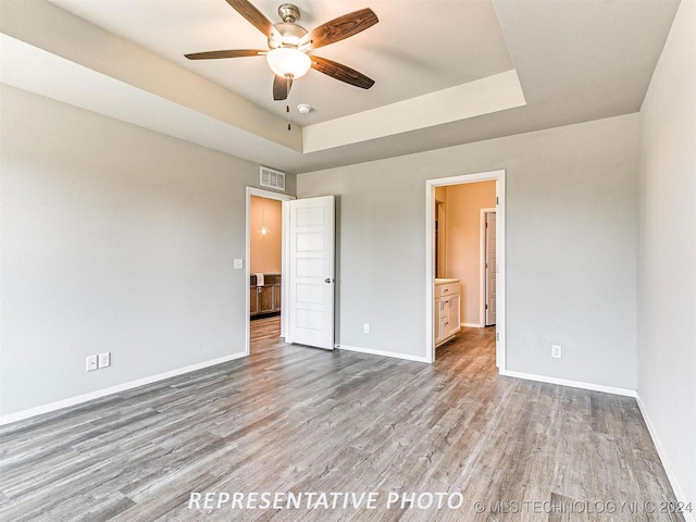 unfurnished bedroom with a tray ceiling, ceiling fan, ensuite bathroom, and wood-type flooring